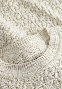 Off-white pleten pulover z teksturiranim vzorcem in rebrastim okroglim izrezom. Oblikovanje vključuje podrobno, tkanino strukturo.