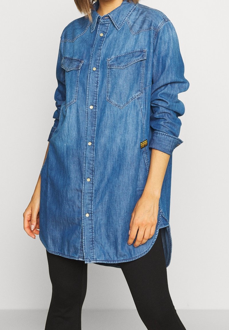 Camicia di denim con bottone frontale, due tasche sul petto e orlo curvo. Colore azzurro chiaro e vestibilità rilassata con maniche arrotolate.