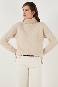 Pulover bej cu guler înalt, cu o croială lejeră, manșete ribbate și un material texturat, purtat cu blugi albi și un curea neagră.