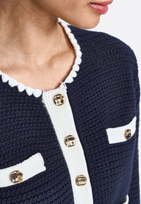 Cardigan lavorato a maglia blu navy con dettagli bianchi, caratterizzato da un colletto tondo a ondine, bottoni dorati e due tasche frontali con strisce orizzontali.