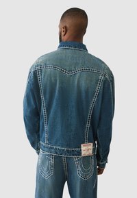 Veste en denim bleu moyen avec une finition texturée. Présente des coutures blanches, un yoke pointu et une étiquette de marque à l'arrière.