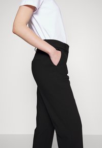 Pantalon noir à coupe ajustée, avec une texture lisse, des poches latérales et des passants de ceinture. Porté avec un t-shirt blanc.