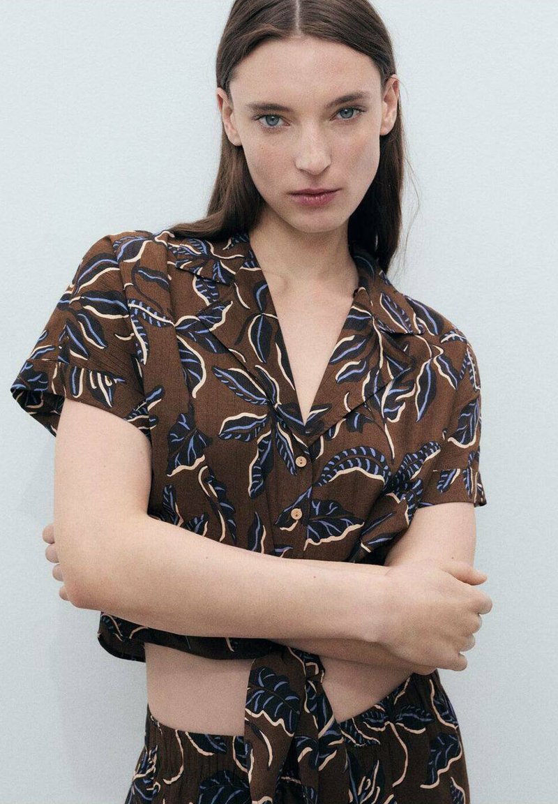 Brown short-sleeve shirt with a collar, featuring a blue and beige leaf pattern, button closure, and tied waist. Cotton-like texture.