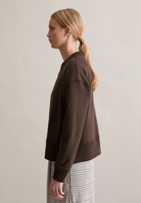 Brązowa bluza z prążkowaną fakturą, okrągłym dekoltem i opuszczonymi szwami na ramionach, zestawiona z kratowaną spódnicą. Widoczne subtelne detale przeszyć.