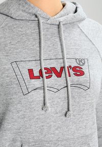Sudadera de algodón gris con un gráfico rojo del logo de Levi's, con capucha con cordón y puños acanalados. Textura suave y diseño casual.