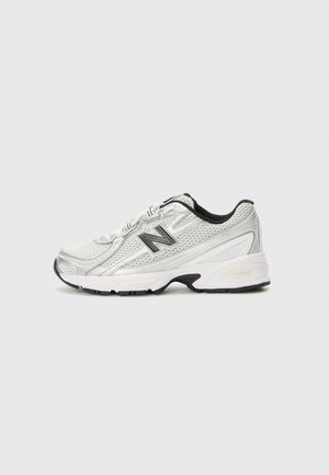 Zapatilla deportiva blanca y plateada con detalles en negro, parte superior de malla, suela gruesa acolchada y un gran logo "N" en el lateral.
