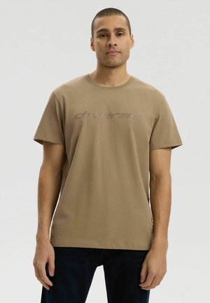 Mężczyzna w prostym brązowym t-shircie z napisem "diverse" na piersi, stojący na neutralnym tle.