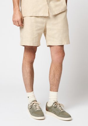 Uomo che indossa pantaloncini beige testurizzati, calzini bianchi alti e sneakers verde oliva con accenti marrone chiaro, in piedi su uno sfondo bianco.