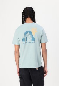 Lyseblå bomull T-skjorte med grafisk design av et kvinnelig ansikt og en sol. Merkenavnet "BILLABONG" er trykket under designet.
