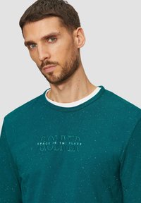 Turkusowy sweter z teksturowanej tkaniny, z okrągłym dekoltem i białymi wykończeniami. Z przodu znajduje się wypukłe logo z napisem "PRZESTRZEŃ JEST MIEJSCEM."