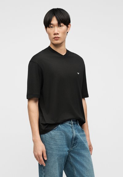 Νεαρός άνδρας που φοράει μαύρη κοντομάνικη μπλούζα V-neck και μπλε τζιν, μπροστά σε ένα απλό ανοιχτό γκρι φόντο.