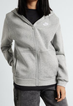 Personne portant un sweat à capuche Nike gris avec fermeture éclair, un T-shirt noir et un jean gris foncé, debout avec une main dans la poche et l'autre main détendue.