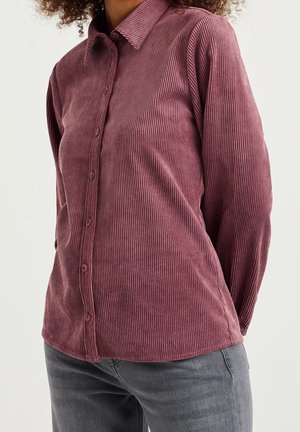Kvinde iført en langærmet, knappet, støvet rosa cordskjorte kombineret med grå jeans, står mod en lys baggrund.