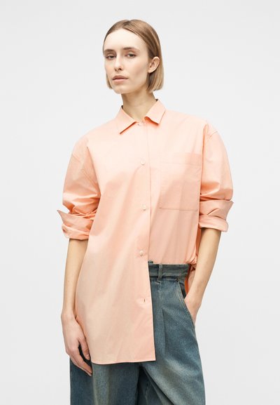 Γυναίκα με κοντά ξανθά μαλλιά, φοράει φαρδιά πουκάμισο σε απαλό ροδακινί χρώμα και φαρδιά μπλε τζιν, με ένα χέρι στην τσέπη, πάνω σε απλό φόντο.