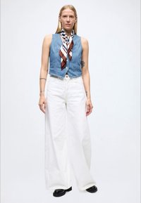 Denimvest med en tettsittende design, kombinert med vide hvite bukser. Et mønstret silkeskjerf i brunt, krem og marineblått fremhever antrekket.