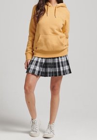 Hoodie giallo in cotone con tasca frontale, abbinato a una gonna plissettata a quadri bianca e nera, completato con sneakers bianche.