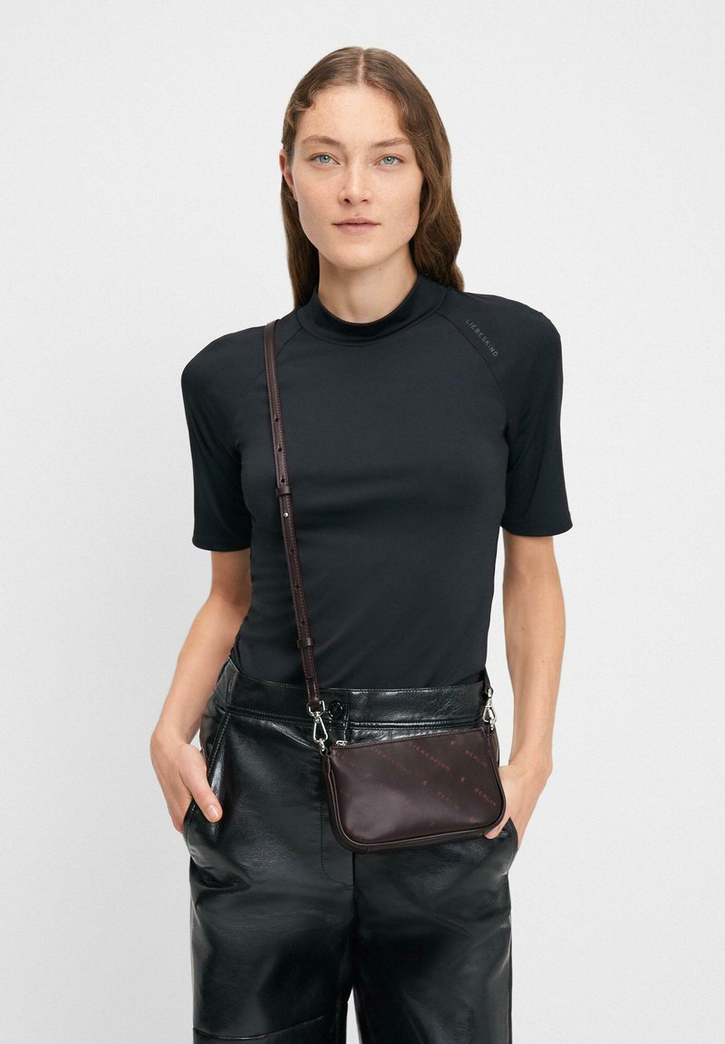 Kobieta o jasnej cerze i falujących brązowych włosach nosi dopasowaną czarną bluzkę z krótkim rękawem, czarne skórzane spodnie oraz małą brązową torebkę na ramię.