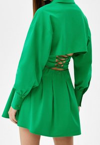 Kobieta w jasnozielonej sukience z długimi rękawami i odkrytymi plecami, zdobionej detalami w formie krzyżujących się sznurowań, stoi na tle białym.