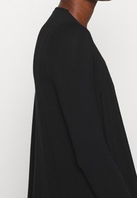 Widok z boku osoby ubranej w czarną, długą koszulę na jednolitym białym tle, ukazujący detale ramienia i górnej części ramienia.