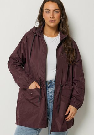 Kobieta ubrana w ciemnofioletową, z kapturem, przeciwdeszczową kurtkę na białej koszuli i niebieskich dżinsach, z jedną ręką w kieszeni kurtki.