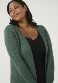 Groene gebreide cardigan met een gemêleerd ontwerp, open voorkant en lange mouwen, gedragen over een zwart top.