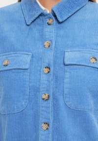 Μπλε καμπαρντίνα πουκάμισο με γιακά, δύο τσέπες στο στήθος και μπεζ κουμπιά. Ο ύφασμα έχει μια υφή με αυλακωτό σχέδιο.