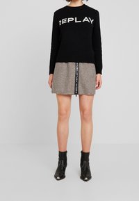 Svart stickad tröja med texten "REPLAY", kombinerad med en brun rutmönstrad kjol med en logotyp och siddragkedja. Svarta ankelstövlar med spetsiga tår.