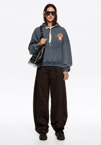 Szara bluza z nadrukiem graficznym, beżowym sznurkiem, zestawiona z luźnymi brązowymi spodniami i czarną teksturowaną torebką. Czarne okulary przeciwsłoneczne i buty.