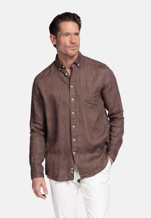 Man with styled dark hair wearing a brown long-sleeve button-up shirt and white pants, standing with one hand in pocket.