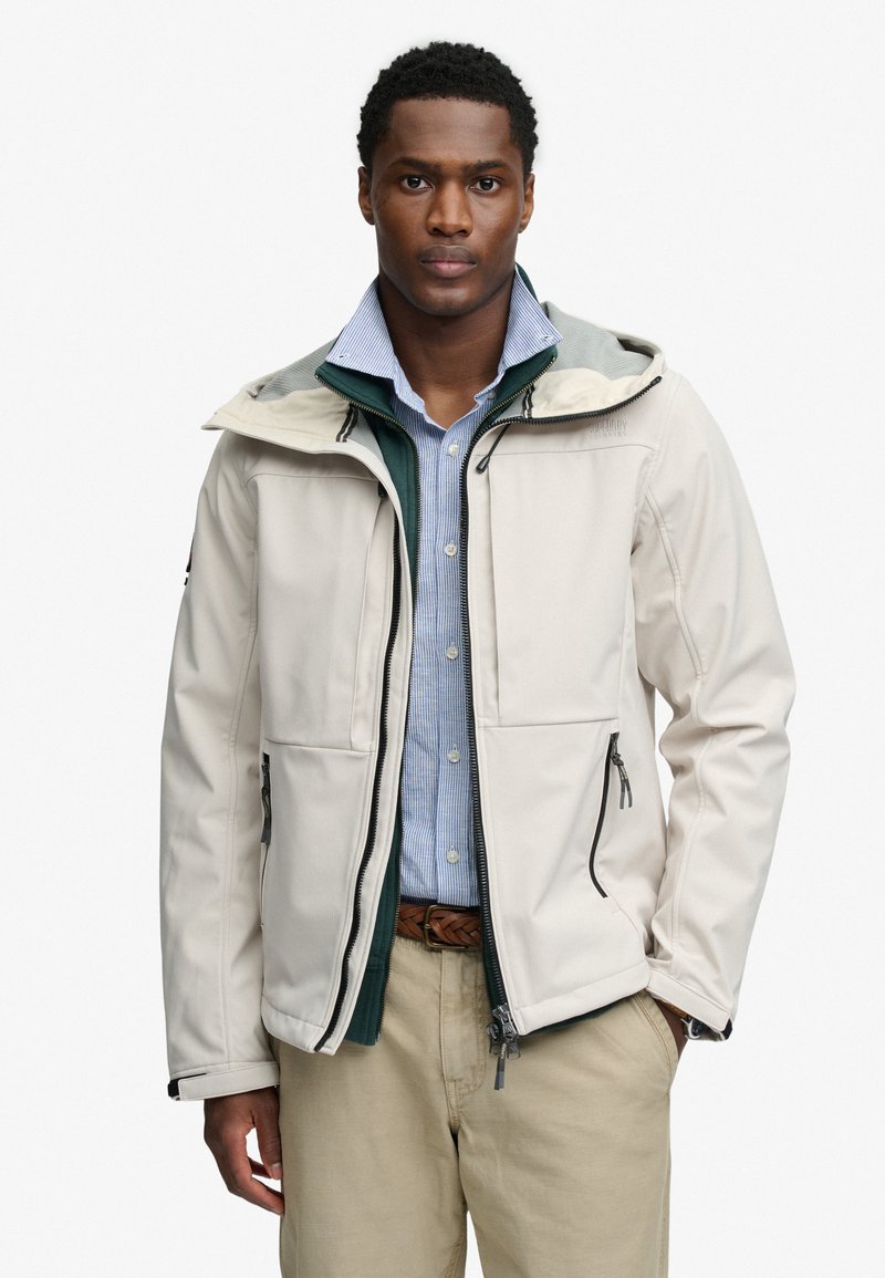 Mann trägt eine hellbeige Kapuzenjacke über einem grünen Zip-up und einem blau gestreiften Hemd mit Knöpfen, dazu Khakihose und geflochtener Gürtel.