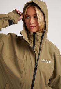 Giacca impermeabile color khaki con colletto alto e cappuccio regolabile. Presenta una cerniera nera e un dettaglio di branding sul petto. Texture liscia.