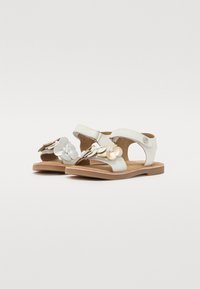 Sandalias de cuero blanco con adornos florales en plata y oro, diseño de punta abierta, correa ajustable y suela marrón.