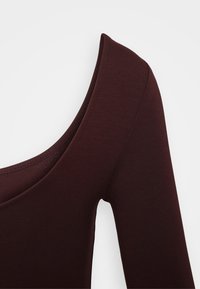 Burgundijska majica dugih rukava s mekanom, rebrastom teksturom i širokim izrezom. Tkanina izgleda rastezljivo i prilagođava se obliku tijela.