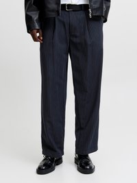 Άντρας που φοράει μαύρο παντελόνι με ρίγες, μαύρη δερμάτινη ζώνη, μαύρα δερμάτινα παπούτσια και μερικώς ορατό μαύρο δερμάτινο μπουφάν.