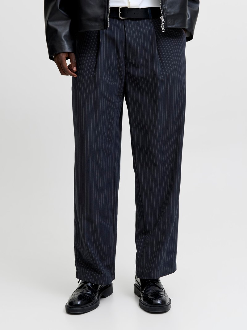 Άντρας που φοράει μαύρο παντελόνι με ρίγες, μαύρη δερμάτινη ζώνη, μαύρα δερμάτινα παπούτσια και μερικώς ορατό μαύρο δερμάτινο μπουφάν.