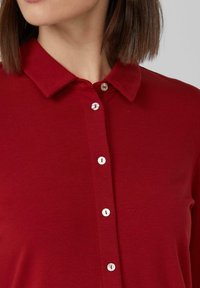 Czerwona koszula zapinana na guziki z klasycznym kołnierzykiem, z jasnymi guzikami i miękką, gładką teksturą. Strukturalny krój i czysty design.