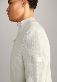 Maglione grigio chiaro a maglia con colletto alto, texture a coste e una toppa con logo sulla manica sinistra. Vestibilità slim, design casual.