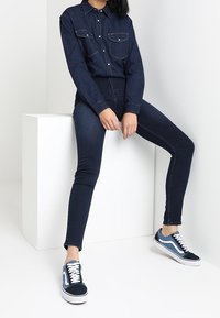 Camisa de mezclilla oscura con botones en la parte delantera y dos bolsillos en el pecho, combinada con jeans ajustados oscuros y zapatillas azules con cordones y acentos blancos.