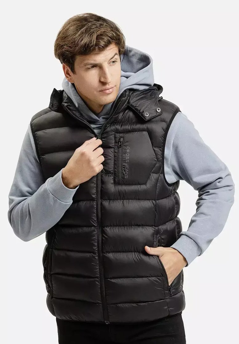 Młody mężczyzna zapinający czarną kamizelkę puchową na szary kaptur, z jedną ręką w kieszeni, patrzący w bok na jasnym jednolitym tle.