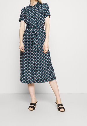 Mujer con vestido midi de manga corta abotonado con patrón geométrico en verde azulado y rosa y sandalias negras con tacón, de pie sobre un fondo blanco.
