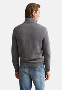 Mann mit dem Rücken zur Kamera, trägt einen grauen, gerippten Rollkragenpullover und hellblaue Jeans vor einfachem weißem Hintergrund.