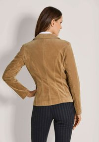 Frau, die ein tailliertes Blazer aus beigem Cord und schwarze Nadelstreifen-Hosen trägt, von hinten vor einem einfarbigen hellen Hintergrund gezeigt.