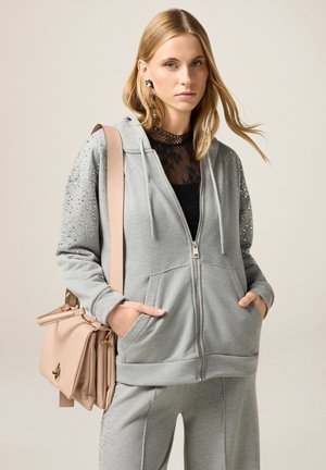 Haut à zip gris avec des accents de strass sur les épaules, en tissu doux, associé à un sac à main rose clair avec des accessoires en or et des zips.