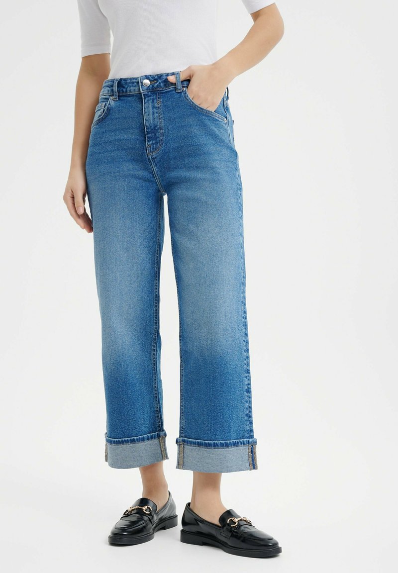 Niebieskie dżinsowe spodnie typu wide-leg z wysokim stanem, z podwiniętym dołem i pięcioma kieszeniami, w zestawie z czarnymi mokasynami.