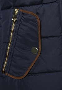 Marineblaue Steppjacke mit einem goldenen Reißverschluss, braunen Wildlederakzenten und einer Druckknopfverschluss-Tasche, die eine gesteppten Textur zeigt.