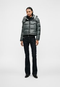 Jachetă padding gri cu fermoar, având un guler structurat și o textură lucioasă, purtată cu blugi evazați din denim închiși la culoare și pantofi negri.