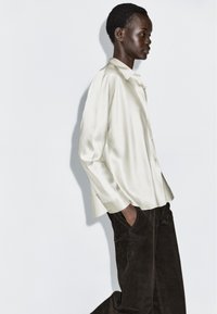 Camicia color crema in satin con bottoni, dalla vestibilità ampia e texture morbida, abbinata a pantaloni di velluto a coste marrone scuro.