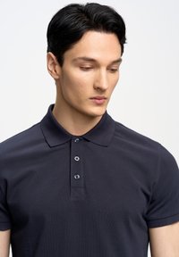 Jūrinės spalvos polo marškiniai, pagaminti iš tekstūruoto audinio su klasikiniu apykakle, turintys tris mygtukus prie apykaklės ir tiesų apatinį kraštą.