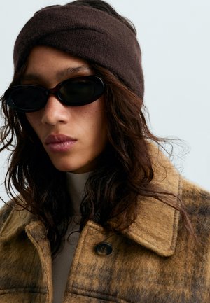 Un modèle aux longs cheveux foncés porte un bandeau en maille marron, des lunettes de soleil noires et un manteau à carreaux sur un fond uni clair.