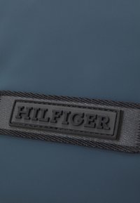 Materiale blu navy con una texture liscia, caratterizzato da un patch in gomma nera con la scritta "HILFIGER" in rilievo su una cinghia intrecciata.